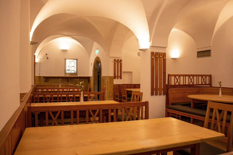 Ein helles Klostergewölbe mit freien Tischen und Stühlen aus Holz.